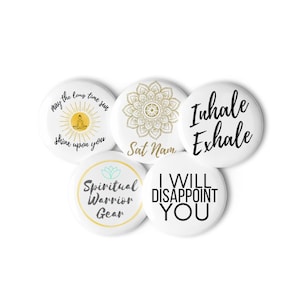May include: A collection of white round buttons with various designs and text. One button features a sun and the words "may the long time sun shine upon you." Another says "Sat Nam," and another says "Inhale Exhale."