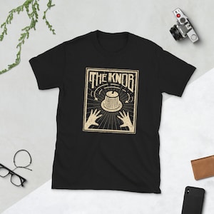 Può includere: T-shirt nera con una grafica in stile vintage di una manopola con il testo "THE KNOB" sopra.