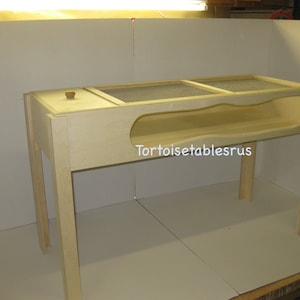 May include: A wooden tortoise table with a mesh top and a hideaway underneath. The table is light brown and has a small wooden knob on the top. The text "Tortoisetablesrus" is visible on the table.