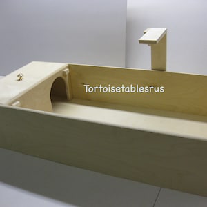 May include: A wooden tortoise habitat with a tunnel, a ramp, and a hinged lid. The habitat is made of natural wood and has a light brown finish. The text "Tortoisetablesrus" is printed on the side of the habitat.
