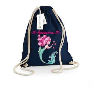 Può includere: Una borsa a cordoncino blu navy con un'illustrazione di sirena e il nome "Ginevra" in lettere rosa. La borsa è realizzata in cotone biologico e ha un cordoncino bianco.
