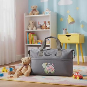 Può includere: Una borsa da viaggio grigia con il nome "Bianca" ricamato sul davanti. La borsa presenta una grafica di tartaruga con corona e fiori. Un orsacchiotto e blocchi di legno sono in primo piano, con un comodino giallo e una libreria sullo sfondo.
