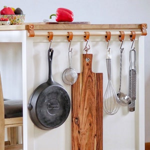 Op de afbeelding: Witte keukeneiland met een houten werkblad en een set haken aan de zijkant. De haken houden een gietijzeren koekenpan, een houten snijplank, een garde, een zeef en een rasp vast. Twee houten stoelen staan voor het eiland.