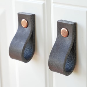 Puede incluir: Dos tiradores de armario de cuero negro con detalles en cobre. Los tiradores están fijados a una puerta de armario blanca.