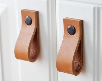 May include: Two tan leather cabinet pulls with a loop design, attached to a white cabinet door. Each pull is secured with a dark, round rivet. The leather has a natural, textured appearance.
