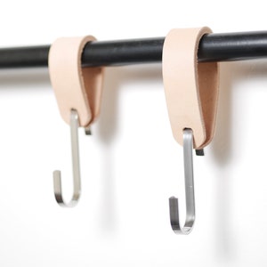 May include: Two natural leather loop hangers with silver metal hooks suspended from a black metal rod. The hooks are designed for hanging items. The background is plain white.
