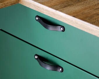 Può includere: Primo piano di un mobile verde con due cassetti. Ogni cassetto ha una maniglia in pelle nera fissata con rivetti argentati. Il mobile è appoggiato su un piano di lavoro in legno.