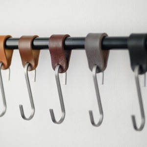 May include: A row of five leather and metal S-hooks in varying shades of brown and black. The hooks are suspended from a black metal rod against a white background. The leather loops are attached to the metal hooks.
