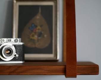 May include: A wooden shelf with a leather strap hanging from it. The shelf is holding a vintage camera and a framed picture with a dried leaf and flowers.