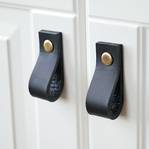 Pode incluir: Dois puxadores de armário de couro preto com botões de metal dourado. Os puxadores estão presos a uma porta de armário branca.