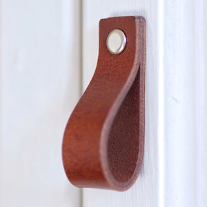 May include: A brown leather cabinet pull with a silver metal screw. The pull is shaped like a loop and is mounted on a white surface.