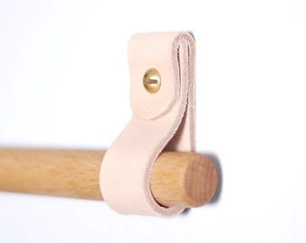 Puede incluir: Una correa de cuero de color claro con un remache de latón está sujeta a una clavija de madera. El cuero está enrollado alrededor de la clavija, creando un diseño simple y minimalista. El fondo es blanco.