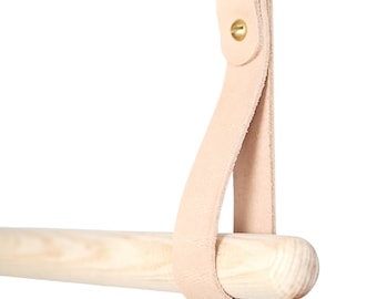 Könnte beinhalten: Eine hellfarbene Handtuchstange aus Holz mit einem Naturlederriemen und Messingbeschlägen. Der Riemen ist mit einer Messingschraube an der Stange befestigt. Die Stange ist zum Aufhängen von Handtüchern oder anderen Gegenständen konzipiert.
