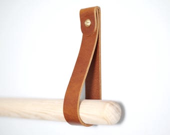 Puede incluir: Un toallero de cuero marrón claro con una barra de madera natural. La correa de cuero está sujeta con un remache de latón. El toallero está diseñado para montarse en la pared.