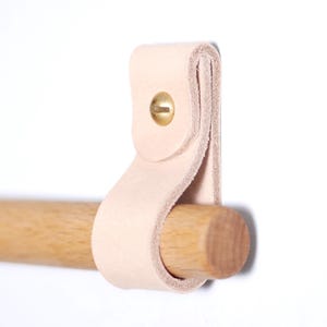 May include: A light-colored leather strap with a brass rivet is attached to a wooden dowel. The leather is looped around the dowel, creating a simple, minimalist design. The dowel is made of light-colored wood.