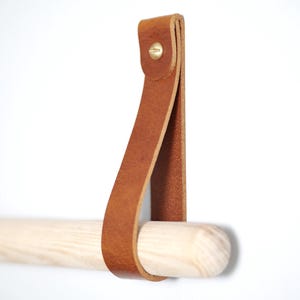 Könnte beinhalten: Ein hellbrauner Lederhandtuchhalter mit einer Holzstange. Der Lederriemen ist mit einer Messingniete befestigt. Die Holzstange ist hell und hat eine glatte Oberfläche. Für die Wandmontage konzipiert, ca. 30 cm lang.