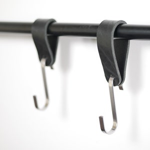 Puede incluir: Dos ganchos de cuero negro con ganchos en S de acero inoxidable colgando de una barra de metal negro. El cuero está doblado sobre la barra. Los ganchos están diseñados para colgar objetos.