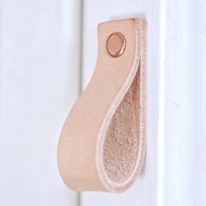 Puede incluir: Un tirador de armario de cuero marrón claro con un tornillo de color cobre. El tirador tiene forma de bucle y una superficie lisa y texturizada.