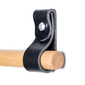 May include: A black leather strap with a silver rivet is attached to a natural wood dowel. The dowel is cylindrical and appears to be made of light-colored wood. The strap is folded over and secured with a metal rivet.