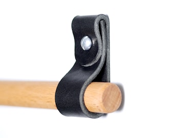 Könnte beinhalten: Ein schwarzer Lederriemen, befestigt mit einer silbernen Niete, hält einen natürlichen Holzdübel. Der Riemen ist um den Dübel geschlungen und erzeugt ein schlichtes Design. Der Hintergrund ist weiß.