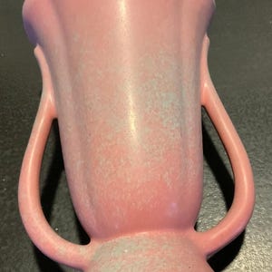 May include: A pink ceramic vase with two curved handles. The vase has a flared base and a ruffled top. The surface has a speckled texture with hints of blue-green. The vase is a decorative item.