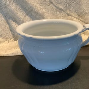May include: A light blue ceramic chamber pot with a handle. The pot has a rounded shape with a wide opening and decorative embossed details. The handle is curved and has a decorative design. The chamber pot is sitting on a dark surface.