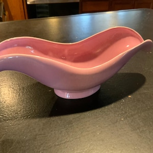 May include: A pink ceramic serving dish shaped like a wave. The dish has a smooth, glossy finish and a rounded base.
