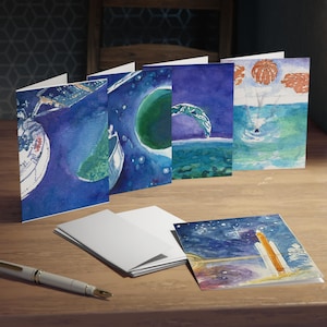May include: A collection of watercolor greeting cards featuring space-themed illustrations. The cards depict planets, rockets, and celestial scenes in shades of blue, green, and orange. A pen and a stack of blank cards are also on the wooden surface.