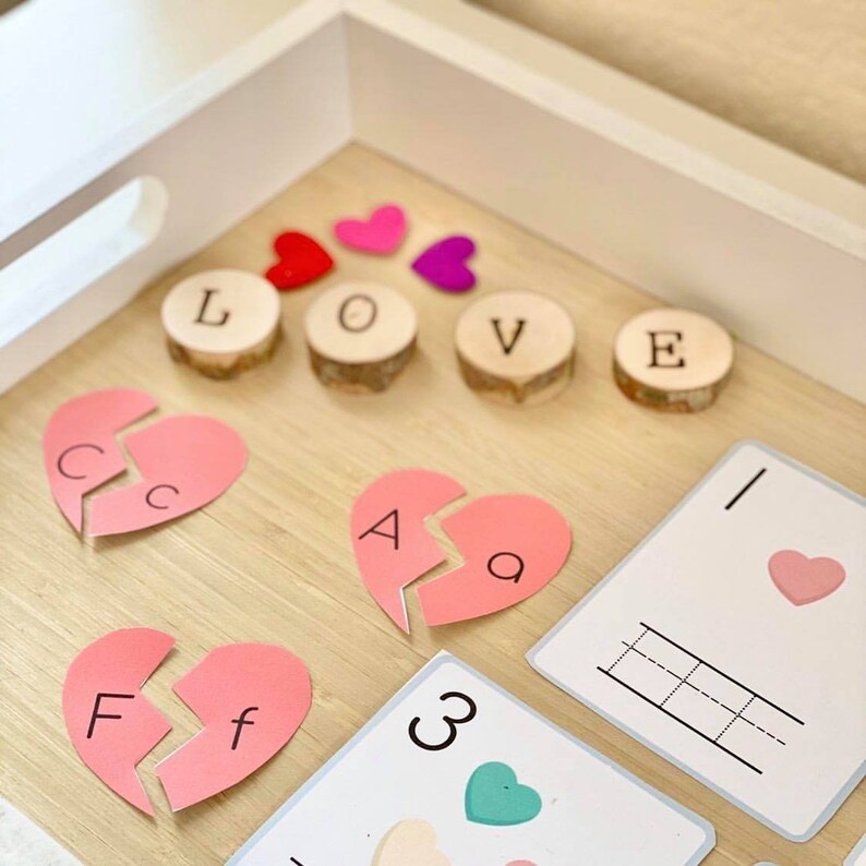 Puede incluir: Una bandeja de madera con corazones de fieltro rosa, fichas de letras de madera que deletrean "LOVE" y corazones de papel rosa con letras en ellos. Tambi&eacute;n hay dos tarjetas blancas con un coraz&oacute;n rosa y una cuadr&iacute;cula en una y un n&uacute;mero 3 y un coraz&oacute;n verde en la otra.
