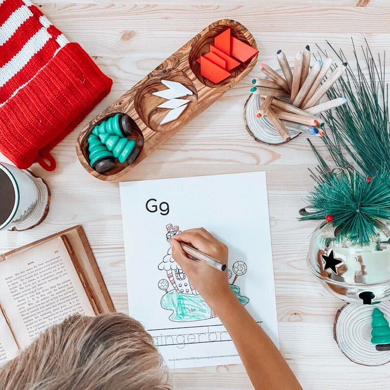 May include: A child is coloring a gingerbread house on a printable worksheet. The worksheet has the letter G and the number 9 printed on it. The child is using a red and white striped crayon. There are other learning materials on the table, including a wooden tray with compartments filled with colorful shapes and a pencil holder with colored pencils.