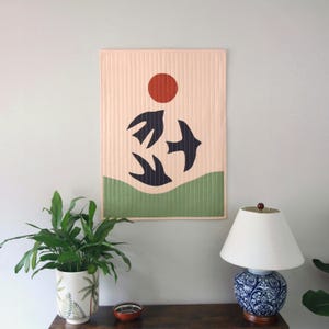 May include: A quilted wall hanging with a minimalist design. The artwork features a red sun, three black bird silhouettes, and a green wavy line on a light pink background. The piece is mounted on a light grey wall.