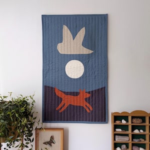 May include: A quilted wall hanging with a nature-inspired design. The artwork features a running fox in orange, a white moon, and a bird-like shape against a blue background. The piece is rectangular and has vertical stitching.