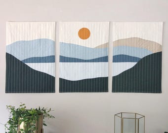 Könnte beinhalten: Drei Wandbehänge mit minimalistischem Landschaftsdesign. Jedes Paneel zeigt eine Berglandschaft in Blautönen, mit weißem Himmel und dunkelgrüner Basis. Das Mittelpaneel enthält eine orangefarbene Sonne. Das Kunstwerk wird an einer weißen Wand ausgestellt.