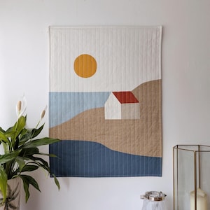May include: A quilted wall hanging depicting a minimalist coastal scene. It features a white house with a red roof, a yellow sun, and blue and brown abstract shapes representing the sea and land. The background is white with vertical stitching.