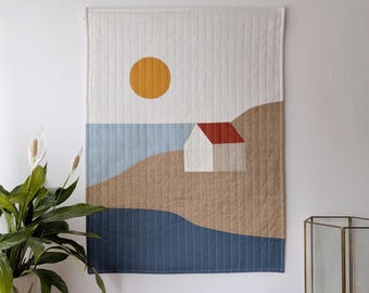 Könnte beinhalten: Eine gesteppte Wandbehänge mit einer minimalistischen Landschaftsszene. Das Design umfasst ein weißes Haus mit rotem Dach, eine gelbe Sonne und blaue und braune abstrakte Formen, die das Meer und das Land darstellen. Der Hintergrund ist weiß mit vertikalen Nähten.