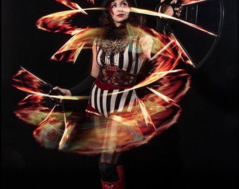 May include: A woman in a black and white striped costume and a top hat performs with glowing LED hoops. The hoops create a swirling pattern of red and orange light against a black background.