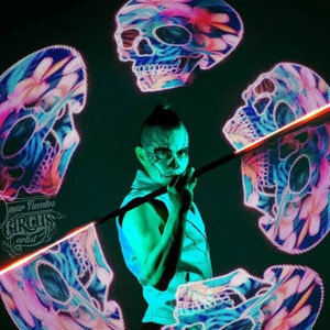 May include: A performer with face paint and a black vest spins a glowing LED staff with skull designs. The staff is lit with red and orange lights. The background is dark with four glowing skull designs in pink, blue, and white.