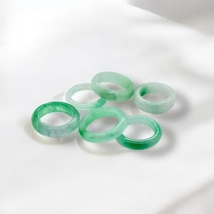May include: A collection of seven jade rings in varying shades of green. The rings are smooth and circular, with a translucent quality. They are arranged on a white surface, with soft shadows.