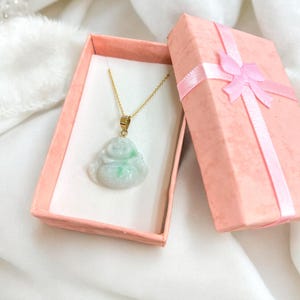 May include: A jade green jadeite pendant in the shape of a sitting Buddha, on a gold chain, in a pink gift box with a pink ribbon.