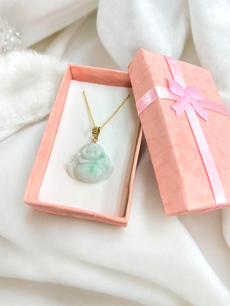 May include: A jade green jadeite pendant in the shape of a sitting Buddha, on a gold chain, in a pink gift box with a pink ribbon.
