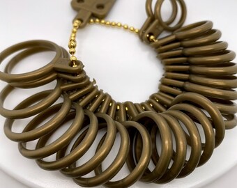 May include: A gold-toned ring sizer with a chain and a small rectangular tag. The sizer has a series of rings of different sizes, all made of a smooth, shiny material.