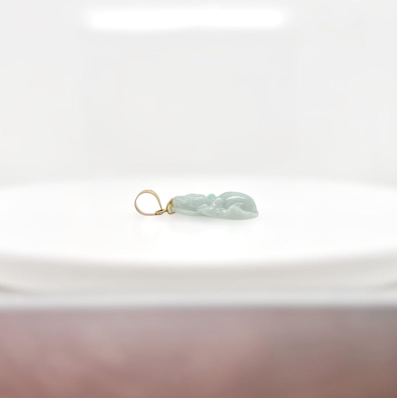 May include: A light green jade pendant carved with a detailed design, set in a gold tone metal bail.