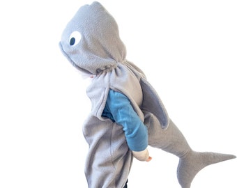 May include: A child wearing a gray shark costume with a white eye detail. The costume has a hood and a tail. The child is wearing black pants and is standing on a white background.