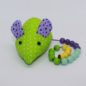 Könnte beinhalten: Eine grüne und violette Stoffmaus mit einem Tupfenmuster und sternförmigen Ohren. Die Maus hat einen Perlen Schwanz mit violetten, gelben und grünen Perlen.