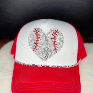 May include: A baseball cap with a red brim and a white crown. The crown features a heart-shaped design made of silver rhinestones with red stitching details. A silver chain accents the brim.