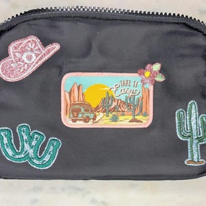May include: Black zippered pouch with embroidered patches. Patches include a pink glitter cowboy hat, a desert scene with a van and the text "Take it Easy", a cactus, and a turquoise design.