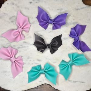 Op de afbeelding: Een verzameling haarstrikken in de vorm van vleermuizen in verschillende kleuren, waaronder roze, paars, zwart en turquoise. De strikken zijn gemaakt van een glad materiaal en liggen op een marmeren oppervlak.