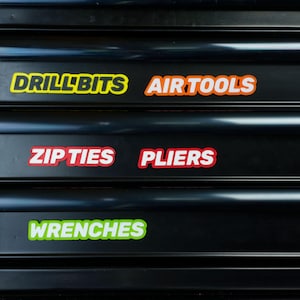 May include: Black tool chest with labels for "DRILL BITS", "AIR TOOLS", "ZIP TIES", "PLIERS", and "WRENCHES". The labels are in various colors, including yellow, orange, red, and green, with white text.
