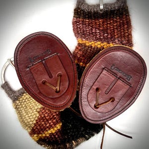 Peut inclure: Deux attaches en cuir marron foncé de forme ovale avec le texte LONCHOP en relief, dotées d'un détail de lacet en cuir central. Elles sont reliées à une sangle textile tissée à rayures horizontales dans des tons marron, beige et noir, avec une boucle en métal argenté.