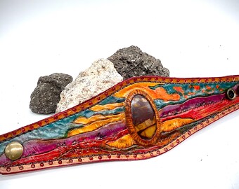 May include: A colorful leather bracelet with a large, oval-shaped stone set in the center. The bracelet is decorated with a multi-colored landscape design and has two metal snaps.